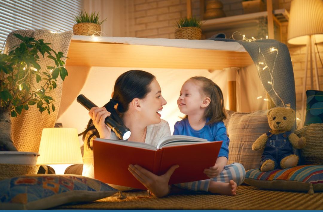 Mãe e filha lendo um livro de capa vermelha dentro de uma cabaninha.