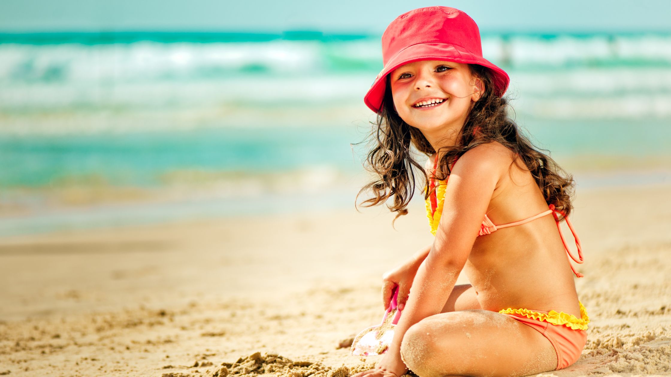 Criança de biquini e chapéu vermelho brincando na areia da praia